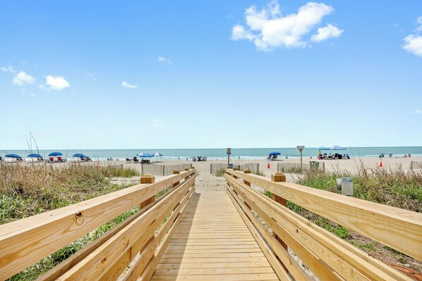 Une promenade en bois menant à une plage où l’on trouve des parasols et des personnes se prélassant au soleil.