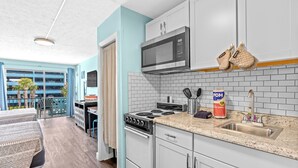 A compact kitchen with a microwave, stove, and sink, a dining area with a table and chairs, and a view of a blue building outside. - Lovely Studio Condo 200 Yards from the Atlantic (Myrtle Beach)