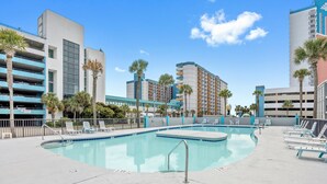 A swimming pool with lounge chairs, palm trees, and modern buildings in the background. - Lovely Studio Condo 200 Yards from the Atlantic (Myrtle Beach)