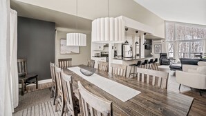 A modern kitchen with a central island, pendant lights, and a dining area with wooden chairs and a large table. - 24-Chalet à Stoneham (Stoneham-et-Tewkesbury)