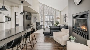 A modern kitchen with a bar area, a fireplace, and a large window with a view of trees. - 24-Chalet à Stoneham (Stoneham-et-Tewkesbury)