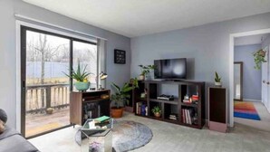 A modern living room with a large sliding glass door leading to a balcony, a flat-screen TV, a bookshelf, and a coffee table.