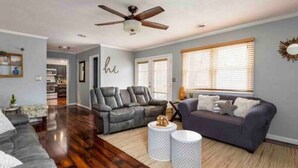A spacious living room with a grey sofa, a smaller grey armchair, and a round coffee table. A ceiling fan is visible, and the room has hardwood flooring.