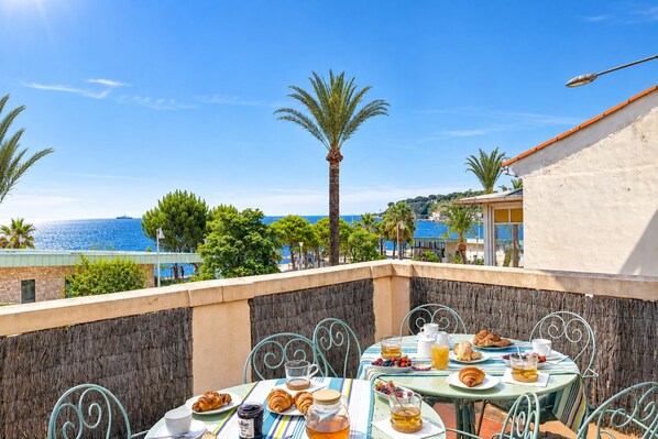 An outdoor dining area with a view of the sea, featuring a table set with croissants, fruits, and beverages.