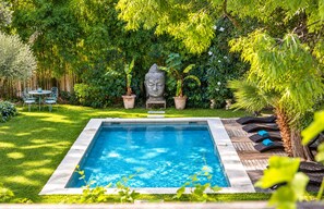 A rectangular swimming pool with a stone border, surrounded by a lush green garden with various plants and a wooden deck with lounge chairs.