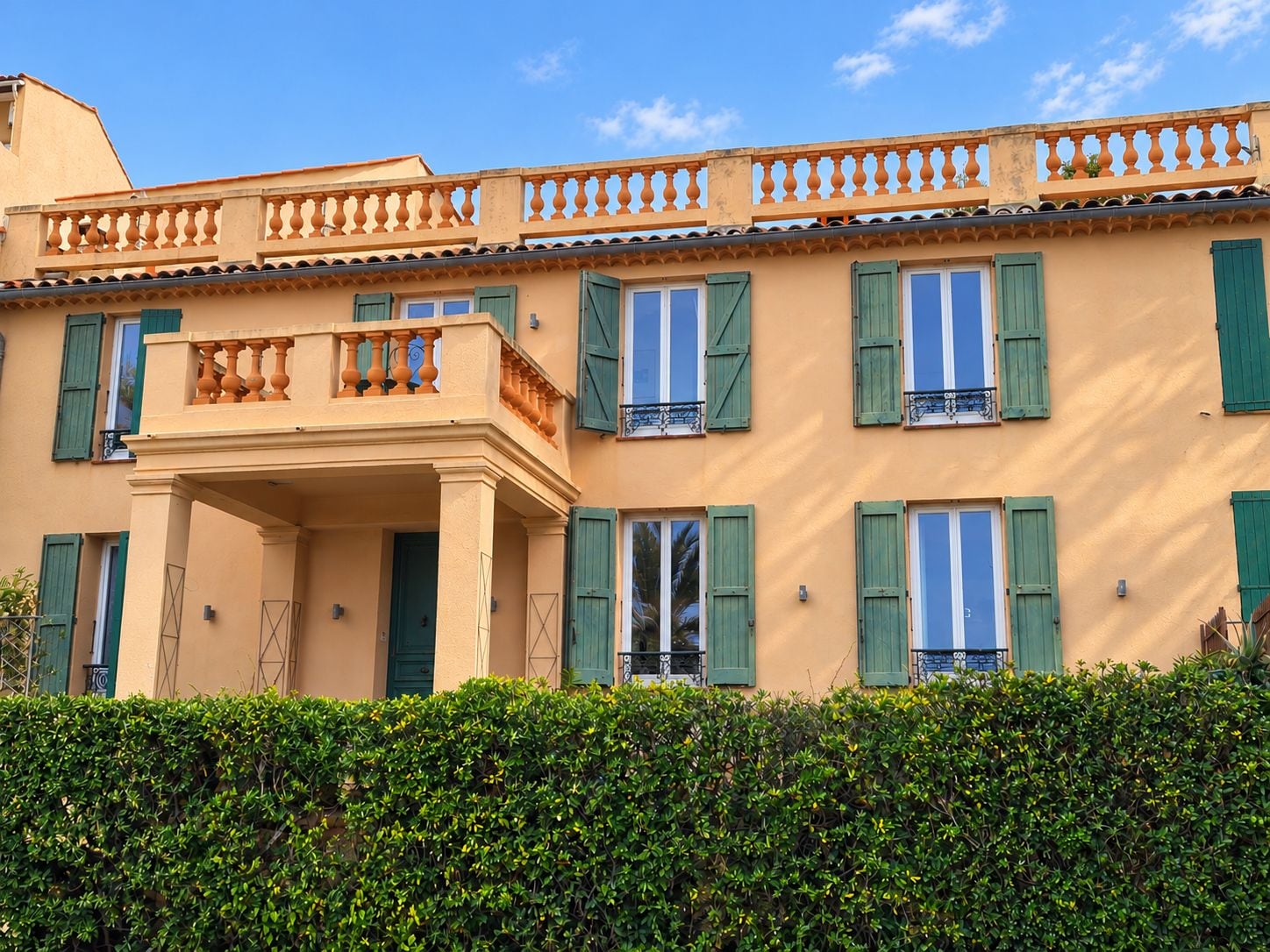 Un immeuble à plusieurs étages doté d’un balcon, de volets verts et d’une haie bien entretenue à l’avant.