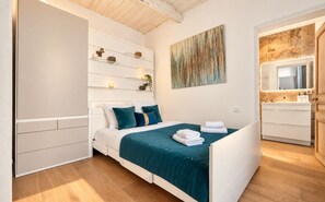 A bedroom with a bed, bedside tables, a wall-mounted shelf with decorative items, a painting, and a bathroom with a mirror and vanity.