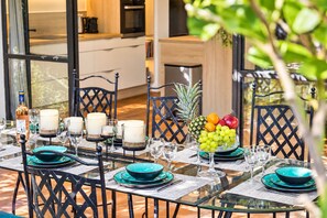 An outdoor dining area with a glass table, black chairs, and a fruit bowl centerpiece.