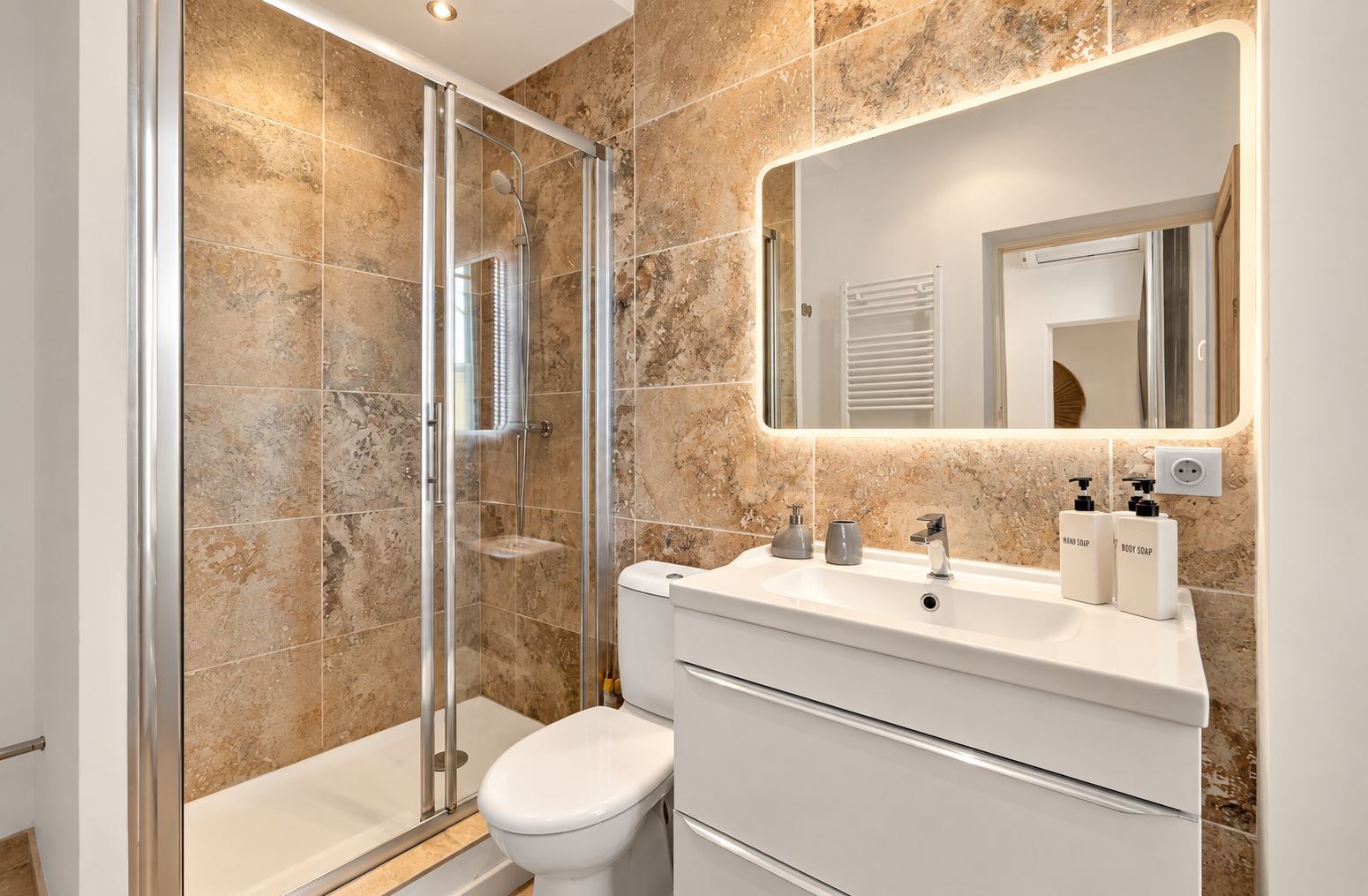 Ein modernes Badezimmer mit einer gläsernen Duschkabine, einer weißen Toilette und einem Waschtisch mit Spiegel und Seifenspendern.