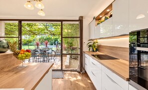 A modern kitchen with a wooden countertop, white cabinets, and a view of a garden through glass doors.