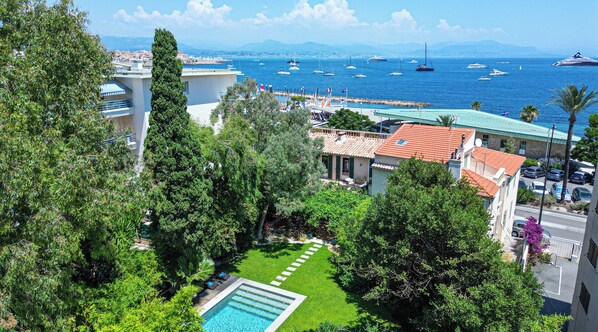 A coastal hotel with a swimming pool, lush greenery, and a view of the sea with boats.