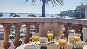 Une table dressée avec des aliments pour le petit-déjeuner et des verres de jus d’orange, donnant sur une plage bordée de palmiers et sur la mer.