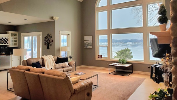 A spacious living room with a large arched window overlooking a body of water, a cozy seating area with a sofa and armchairs, and a kitchen area with cabinets and a dining table. - Lakefront Sunsets and Sandy Beach in Rockford, MI (ROCKFORD)