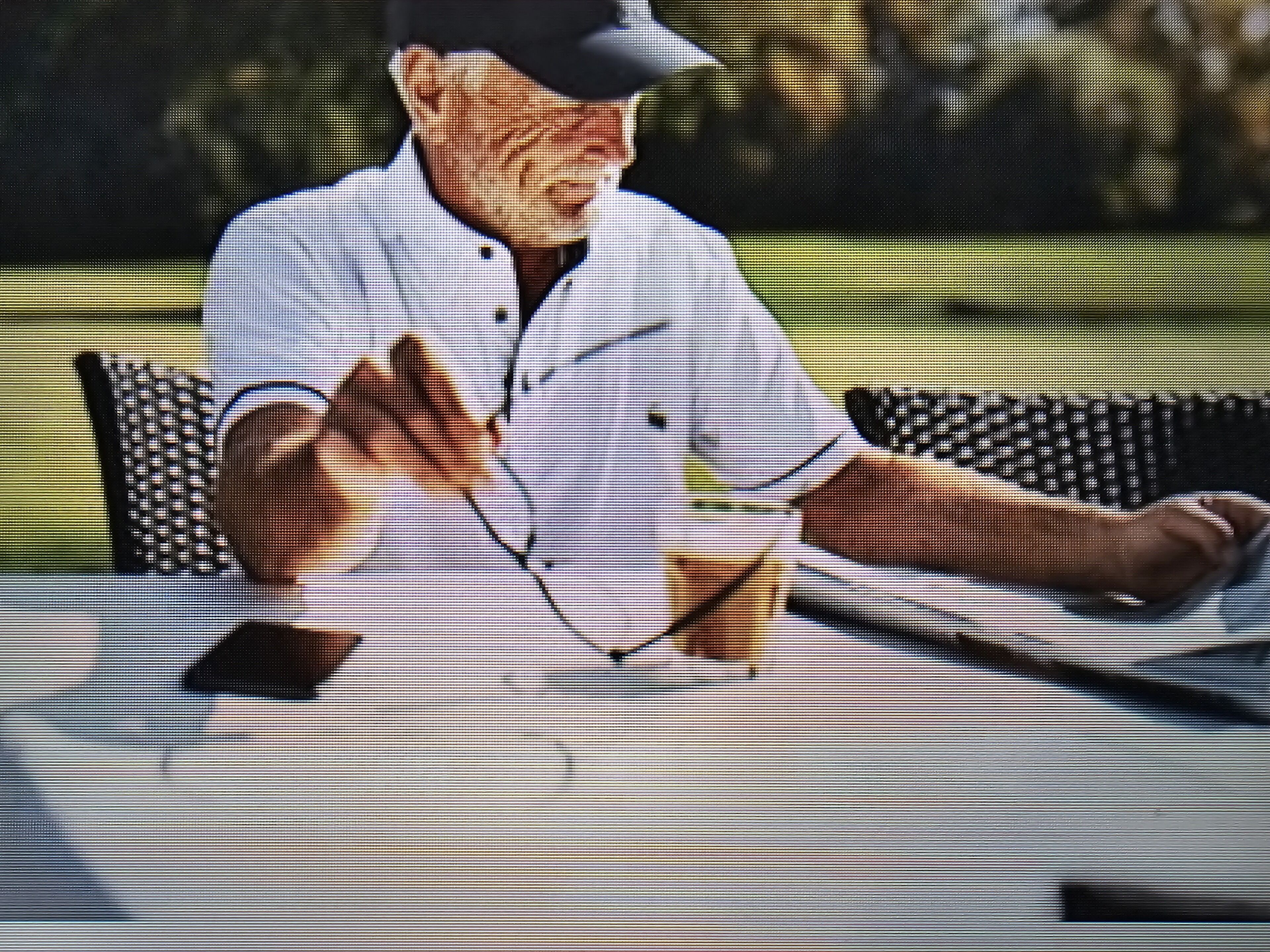 A person sitting at a table with a drink, wearing a white shirt and a cap.