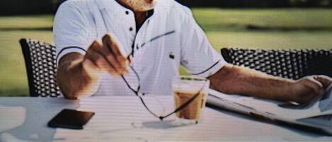 Une personne assise à une table, un verre à la main, vêtue d’une chemise blanche et d’une casquette.