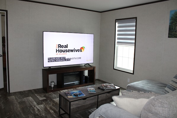 Ein Wohnzimmer mit Fernseher, der 'Real Housewives of Atlanta' zeigt, einem Sofa, einem Couchtisch mit Zeitschriften und einem Fenster mit Jalousien.