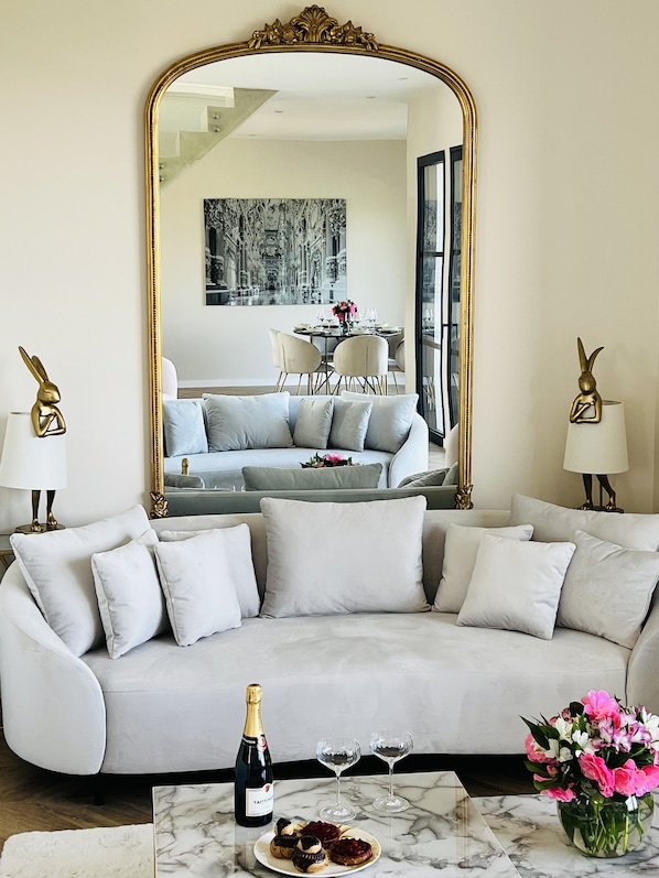 A modern living room with a large mirror, a sofa, a coffee table with a bottle and glasses, and a vase of flowers.
