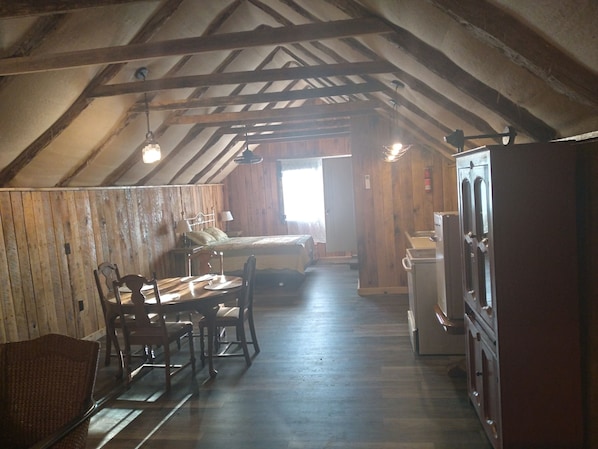 A rustic attic bedroom with a wooden ceiling, a bed, a dining table with chairs, and a kitchenette.