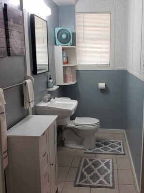 Et lille badeværelse med toilet, håndvask og underskab. Der er to tæpper på gulvet, en ventilator på en hylde og et vindue med rullegardiner.