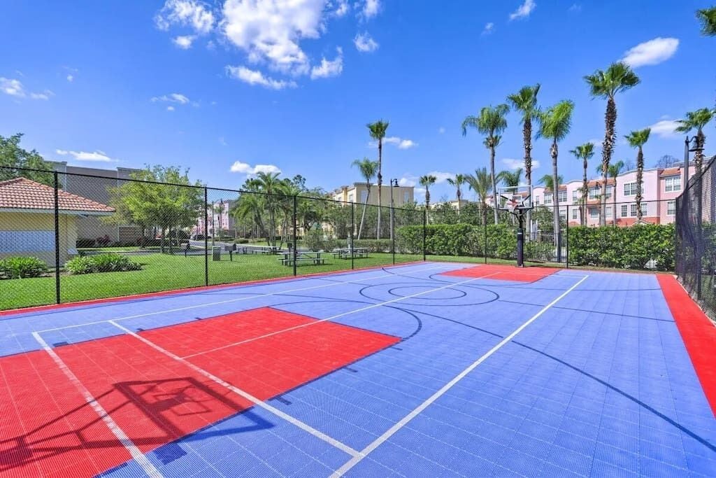 Een buitenbasketbalveld met rode en blauwe markeringen, omgeven door een hek en palmbomen.