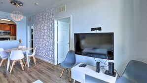 Uma sala de estar moderna com uma televisão de tela plana, uma área de jantar com uma mesa e cadeiras brancas, e uma cozinha visível por uma porta aberta.