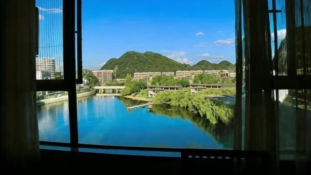 A view through a window of a building overlooking a river, with greenery and distant hills. - Longhuyuan Hostel (Yanshan)