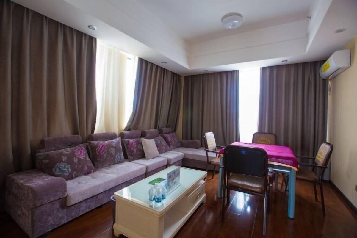 Una sala de estar con un sofá modular, una mesa de centro, una mesa de comedor con sillas y ventanas grandes con cortinas.