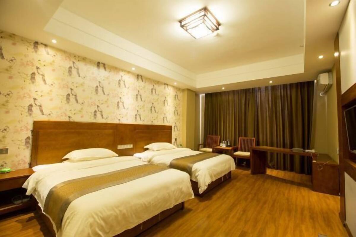 Habitación de hotel con dos camas, suelo de madera, un ventanal grande con cortinas y un televisor.