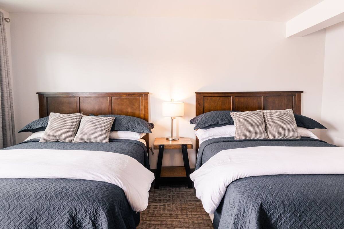 A hotel room with two beds, each with a wooden headboard, a nightstand between them, and a lamp on the nightstand.