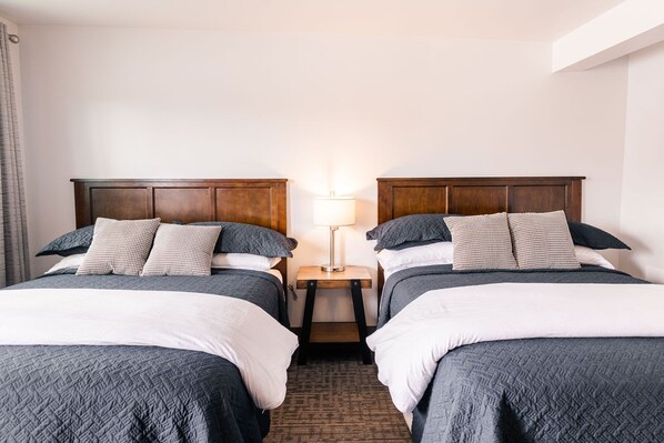 A hotel room with two beds, each with a wooden headboard, a nightstand between them, and a lamp on the nightstand. - Alaska Frontier Inn (Delta Junction)