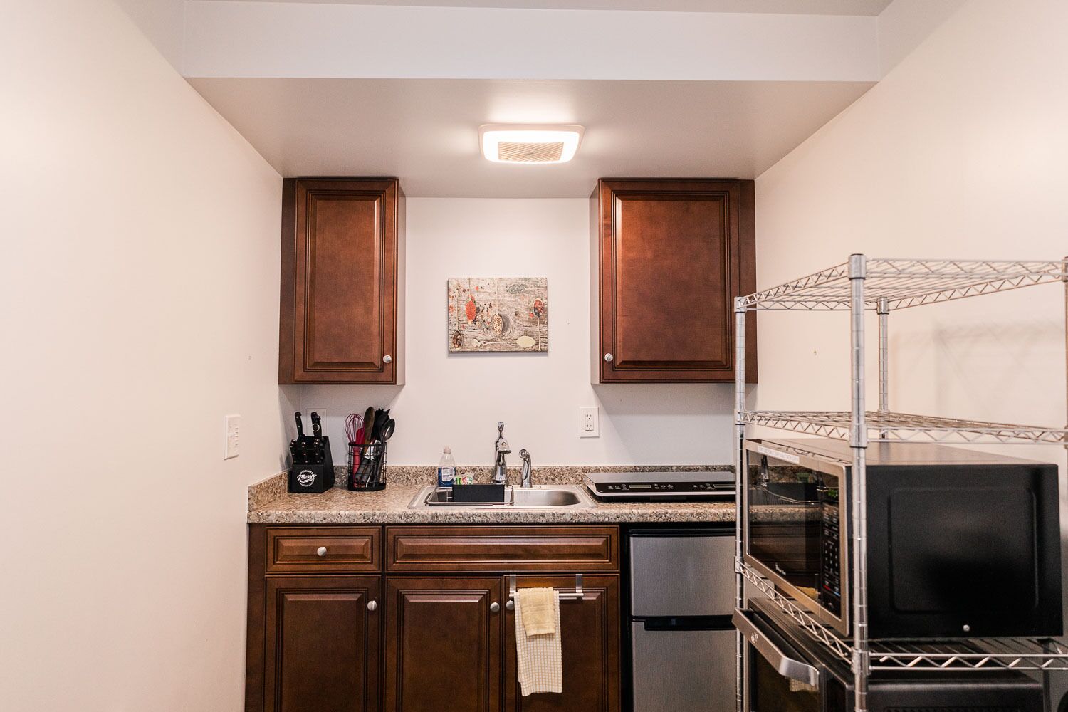 A compact kitchen with wooden cabinets, a stainless steel refrigerator, a built-in oven, and a metal rack.