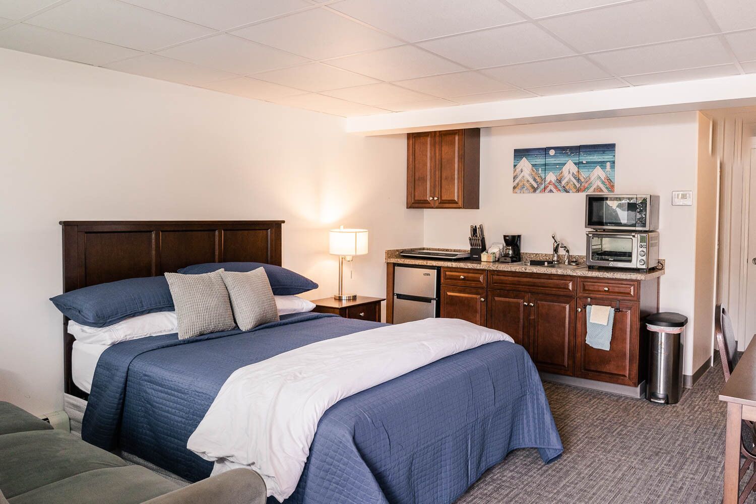A hotel room with a large bed, a kitchenette, a small trash can, and a painting on the wall.