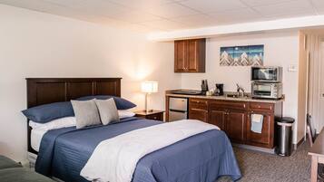 A hotel room with a large bed, a kitchenette, a small trash can, and a painting on the wall.