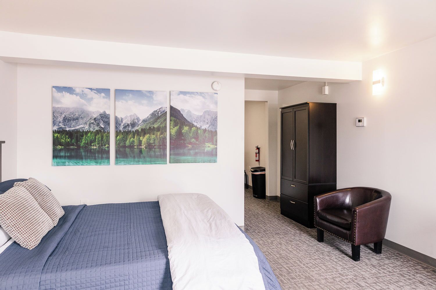A hotel room with a bed, a chair, and three framed pictures of mountains and lakes on the wall.