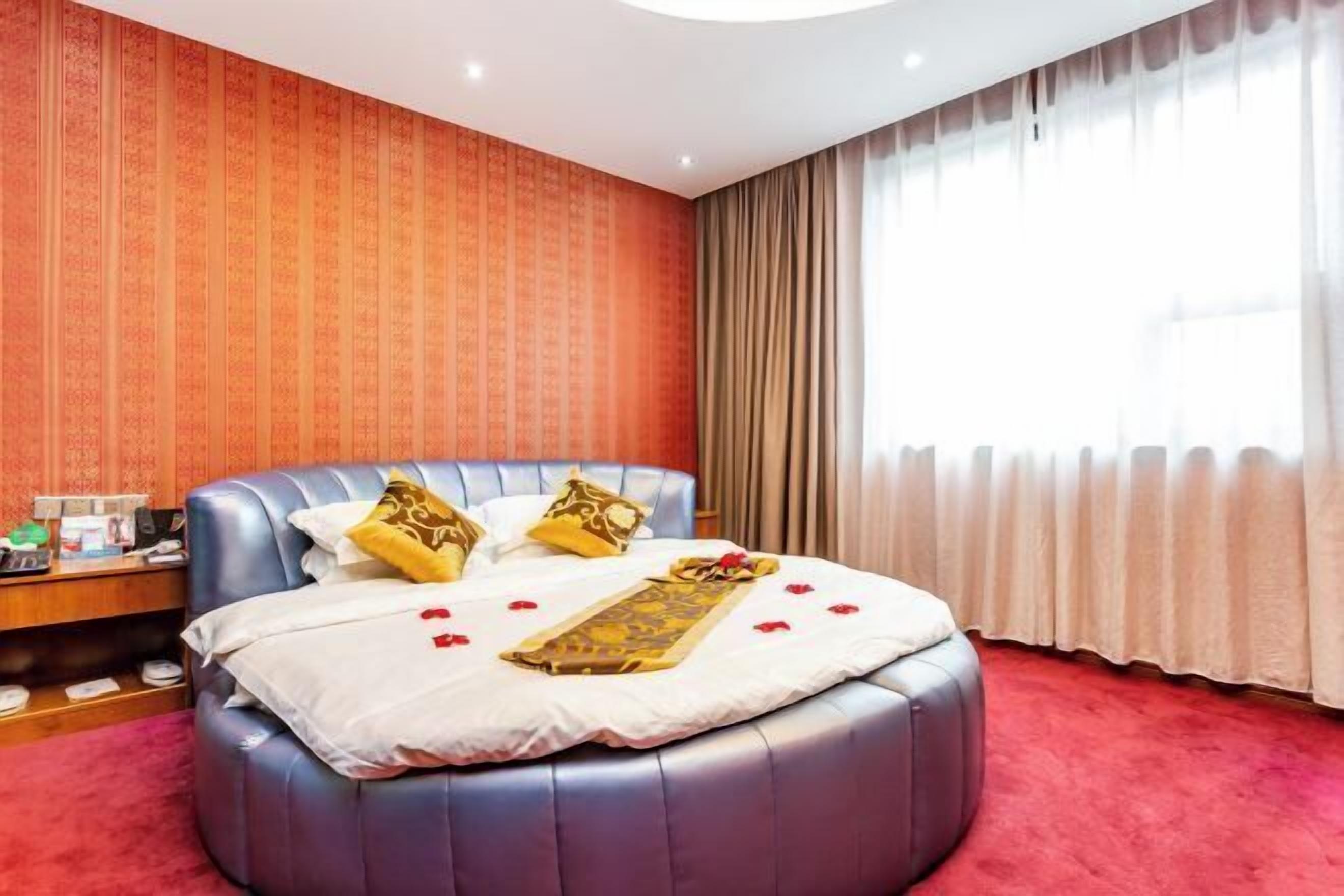 A room with a curved bed, a nightstand, a lamp, and a window with curtains.