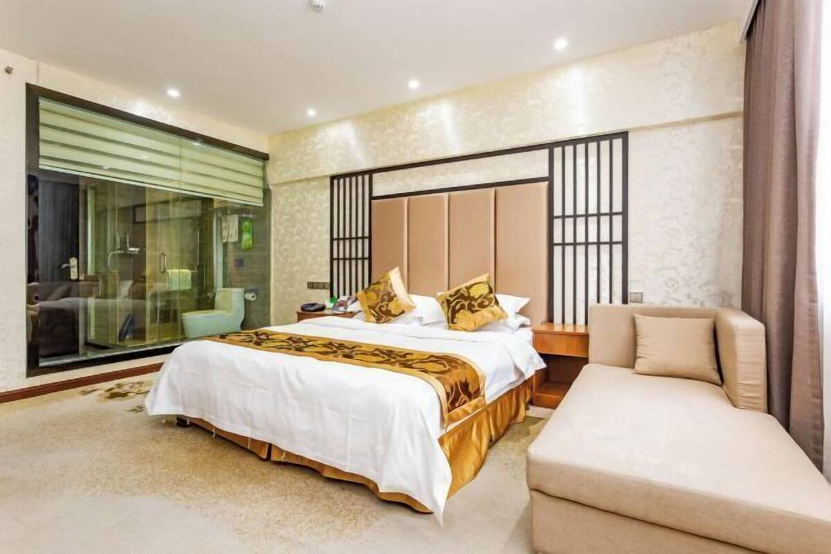 A bedroom with a bed, a sofa, a glass-enclosed bathroom, and a large window.