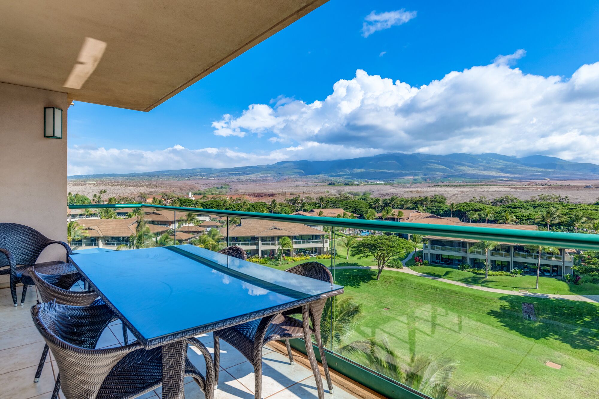 A balcony with a glass table and wicker chairs, overlooking a well-maintained lawn and distant mountains.