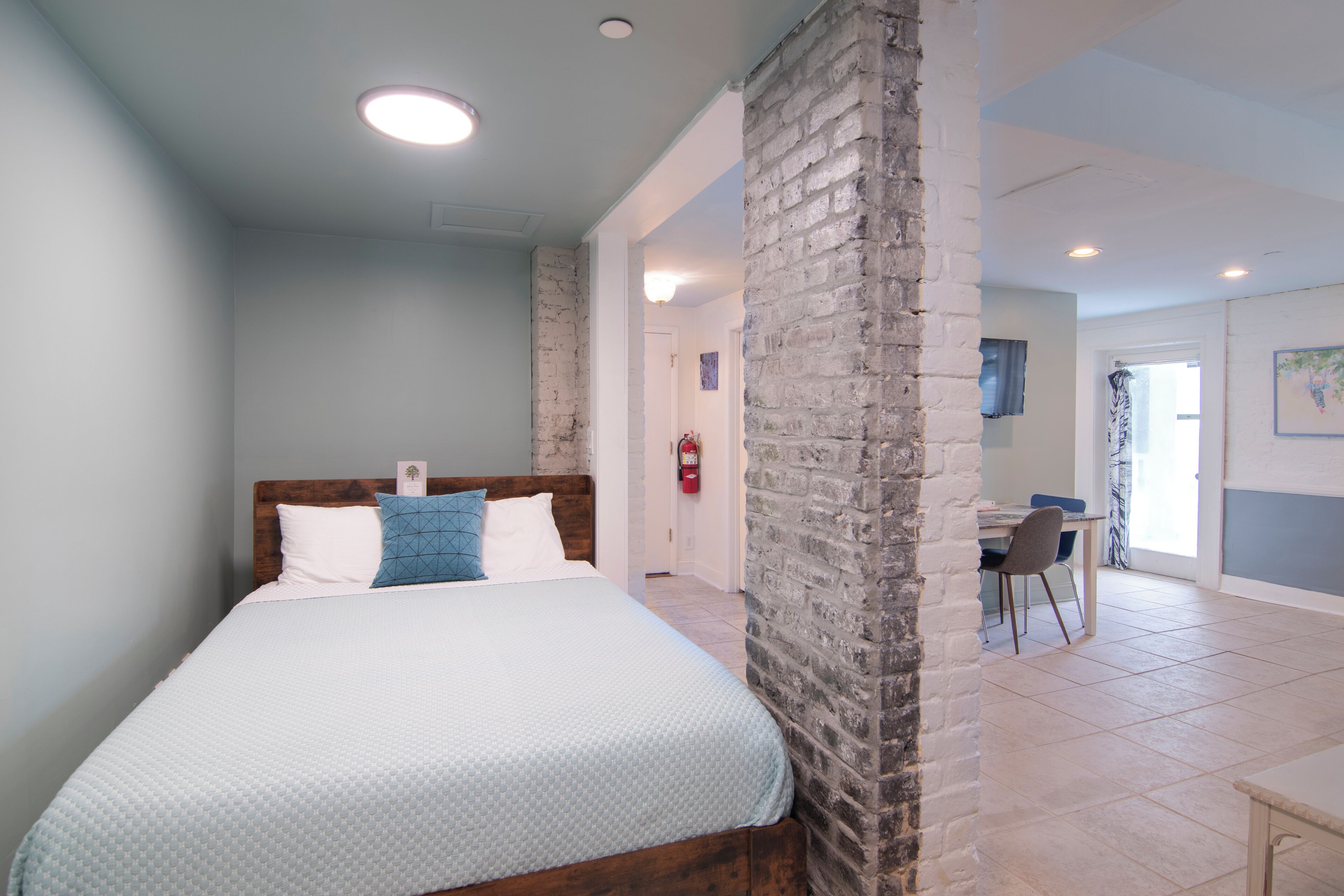 Un dormitorio con cama, pared de ladrillo decorativa, escritorio con silla y puerta que da a otra habitación.