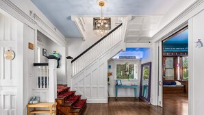 A grand staircase with white paneling and a chandelier, leading to a room with a blue accent wall and a view of the outdoors. - Tree House (Birmingham)