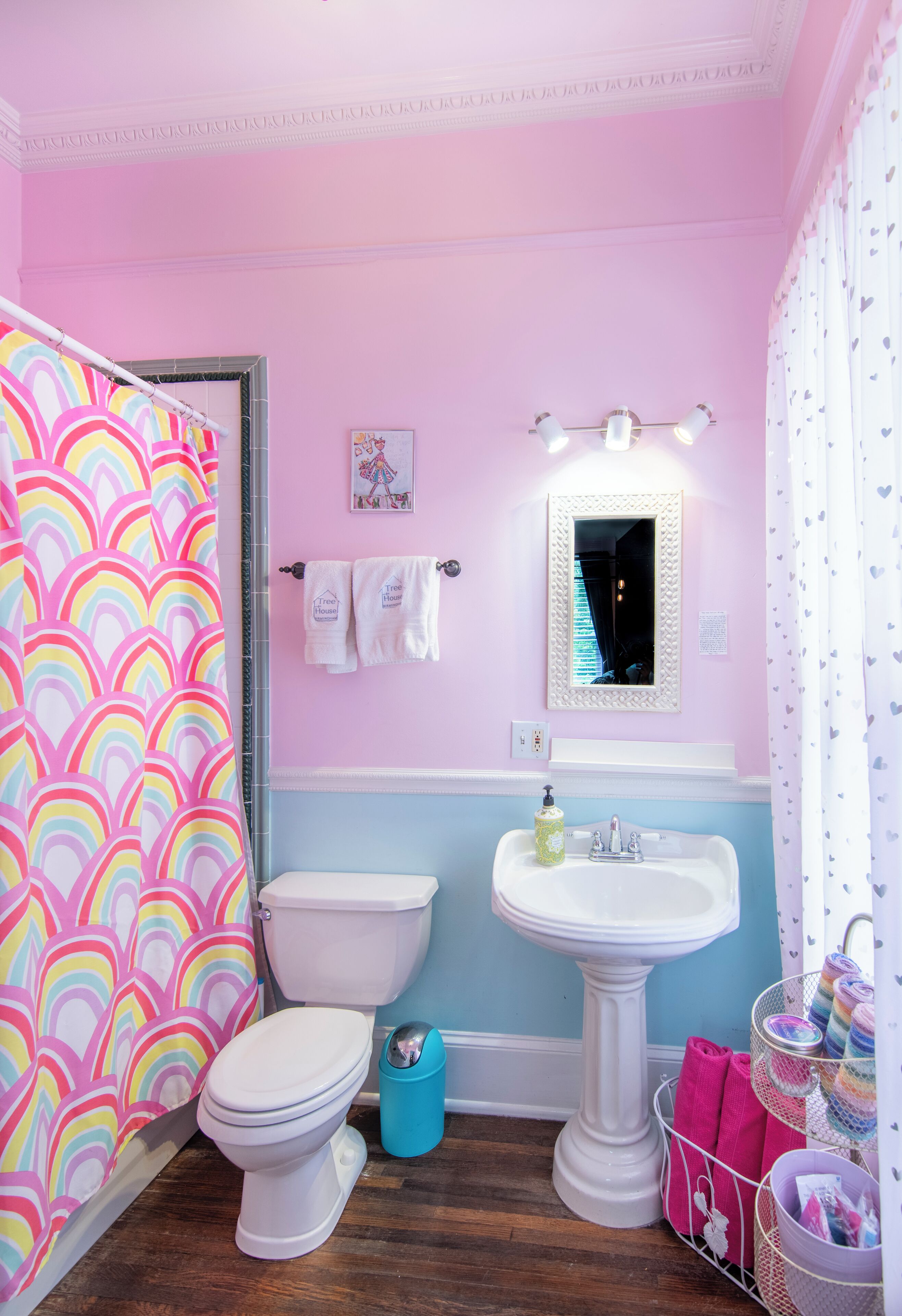 Ein Badezimmer in Rosa und Weiß mit Toilette, Waschbecken auf Säule und einer Duschvorhang mit Regenbogenmuster.