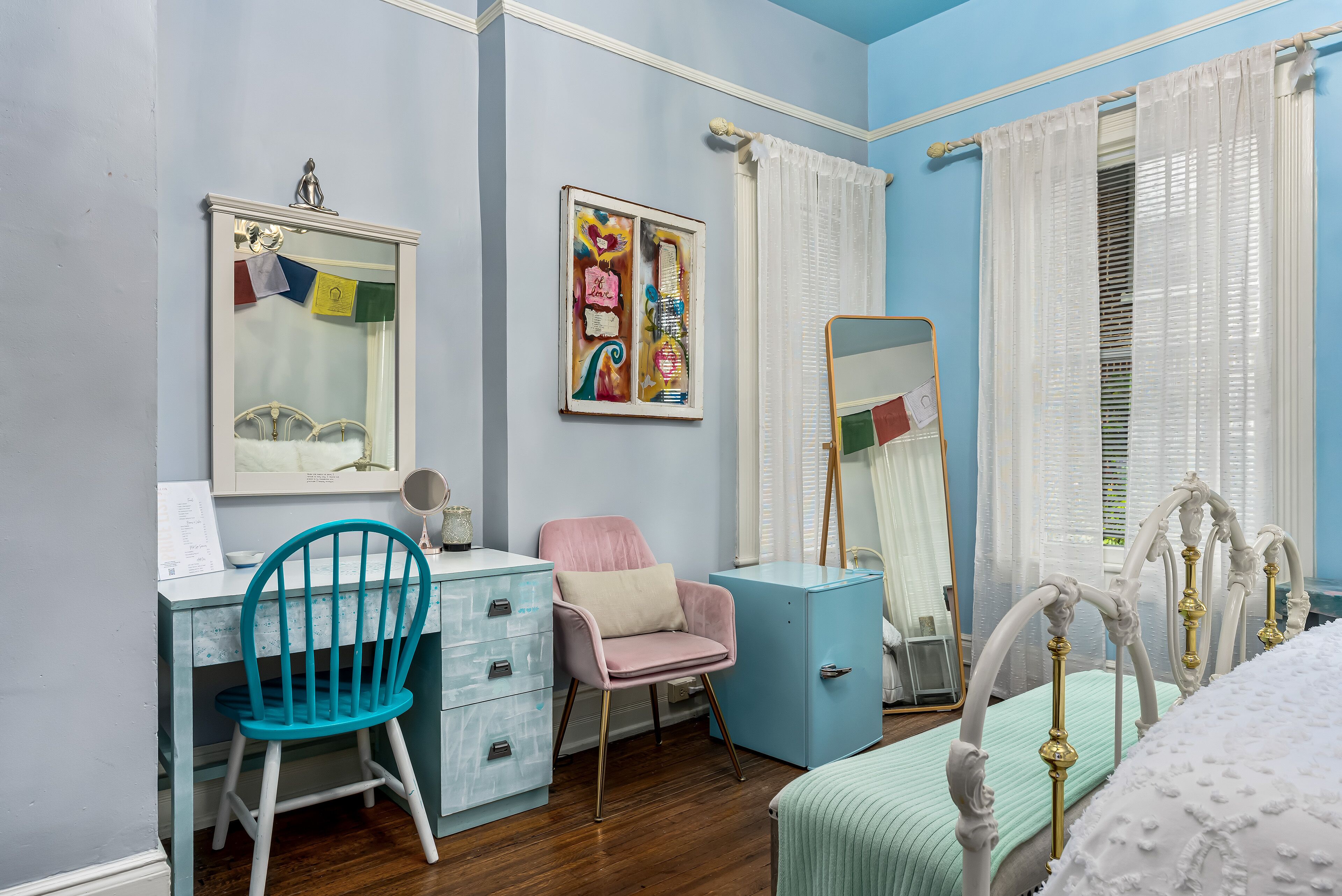 Une chambre avec une commode bleue, une chaise rose, un lit avec une tête de lit blanche et un miroir.