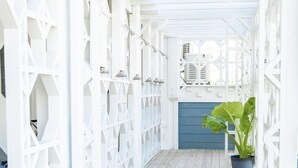 Um corredor de madeira clara, com uma porta azul no final, que leva a uma área coberta com teto branco e uma unidade de ventilação.