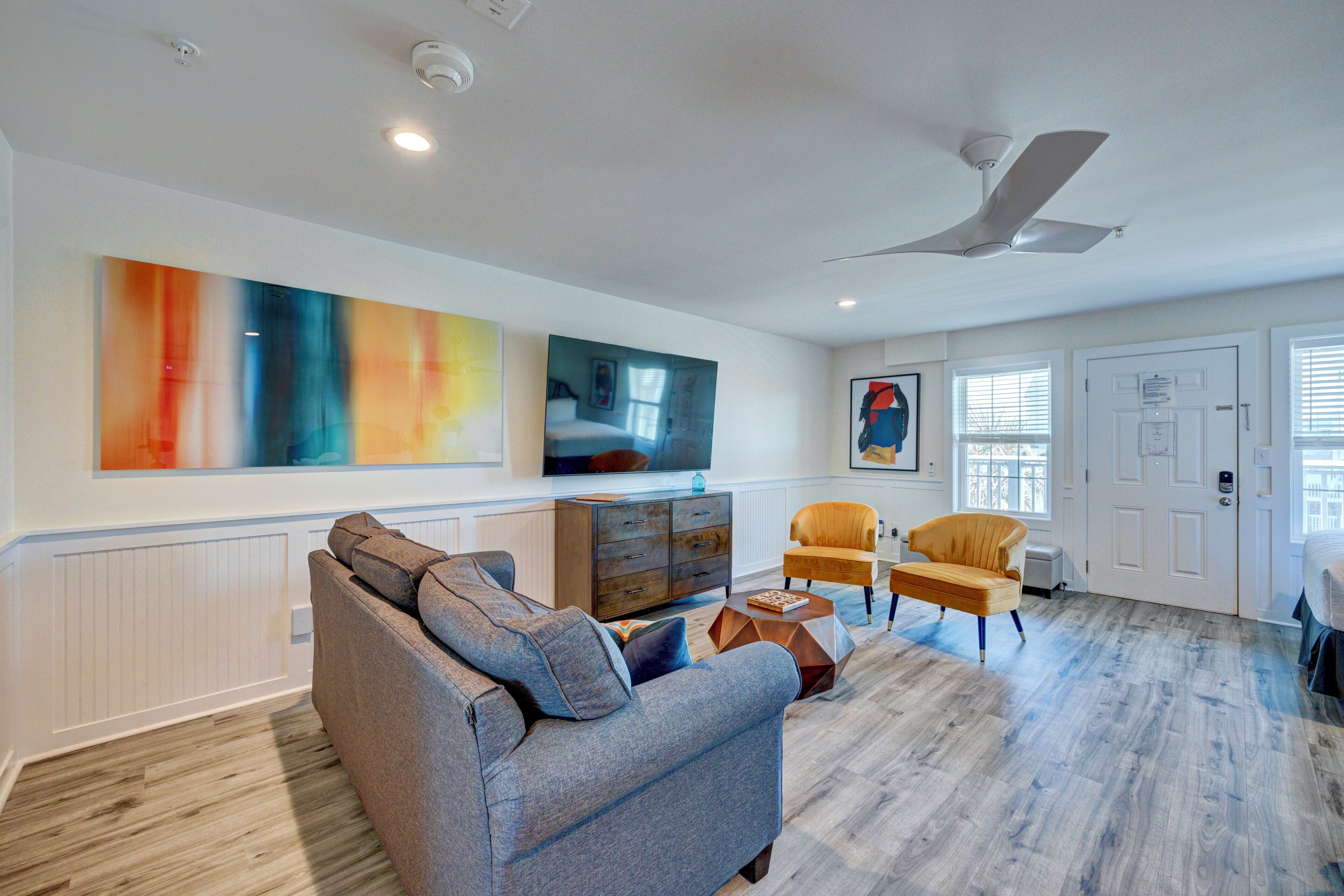 A modern living room with a grey sofa, a wooden coffee table, and a yellow armchair. There is a flat-screen TV mounted on the wall, a ceiling fan, and a large abstract painting.