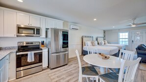 Une cuisine moderne équipée d’appareils électroménagers en acier inoxydable, un coin repas avec des chaises blanches et une chambre avec un lit et une fenêtre.