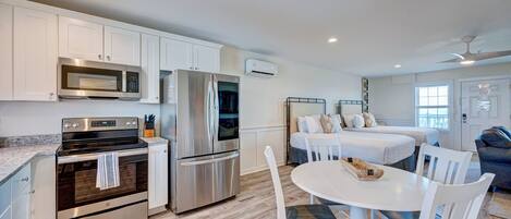 A modern kitchen with stainless steel appliances, a dining area with white chairs, and a bedroom with a bed and a window.
