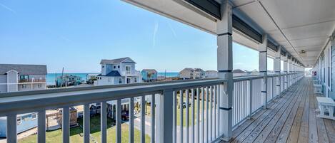 A covered wooden deck with white railings, overlooking a beach and houses.