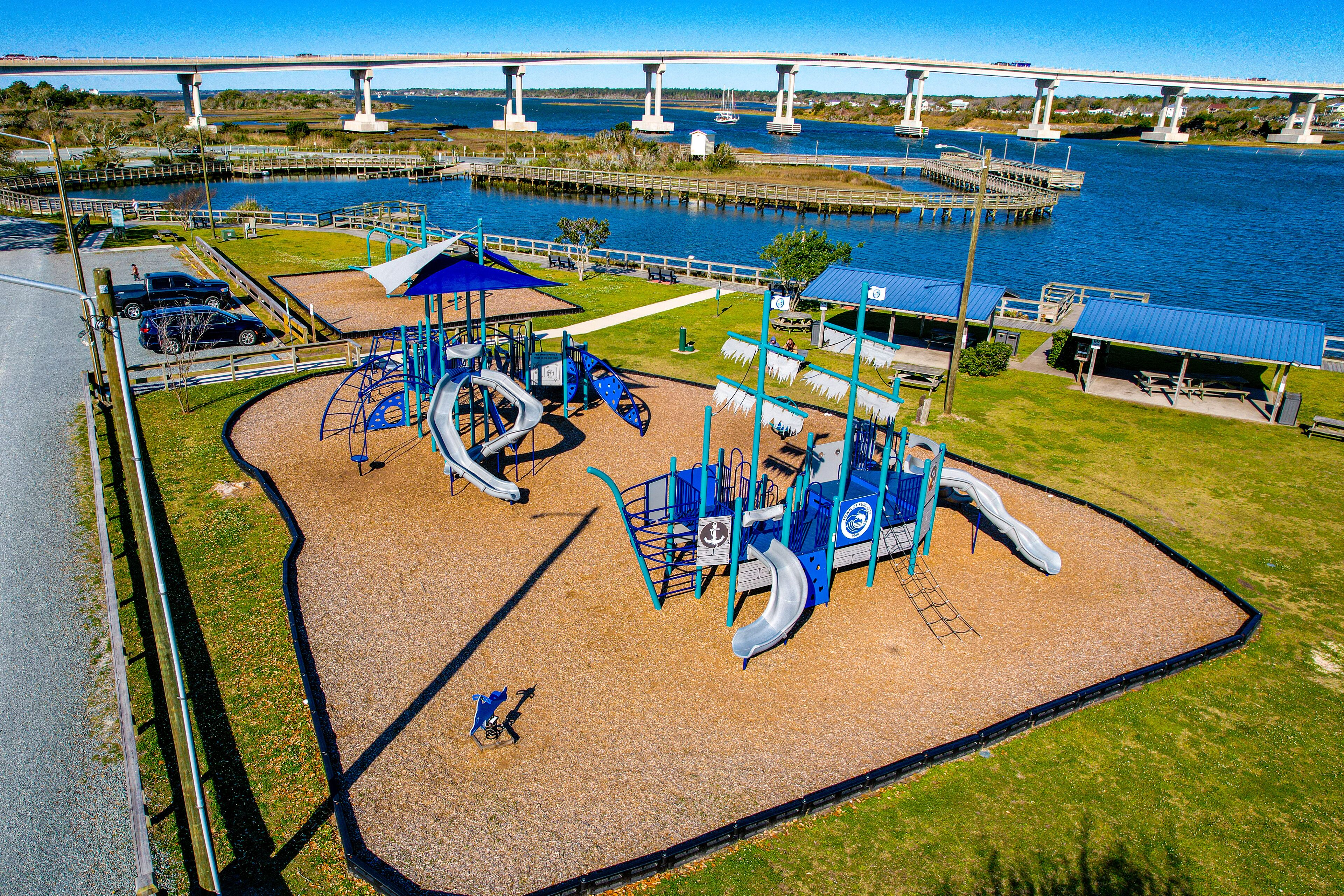 A playground with slides and swings, a picnic area, and a view of a bridge over water.