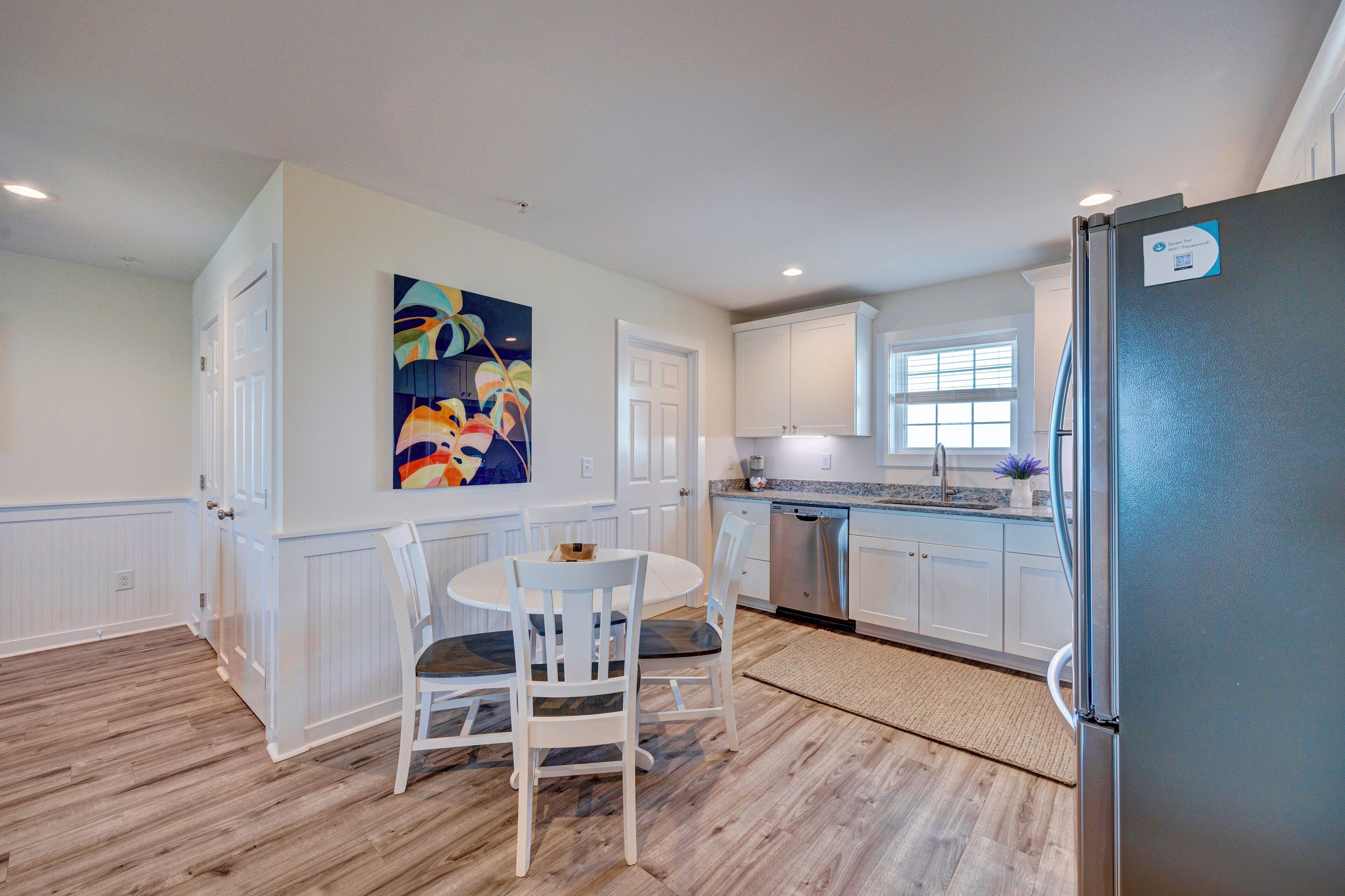 A kitchen with white cabinetry, a stainless steel refrigerator, a dining table with chairs, and a colorful painting on the wall.