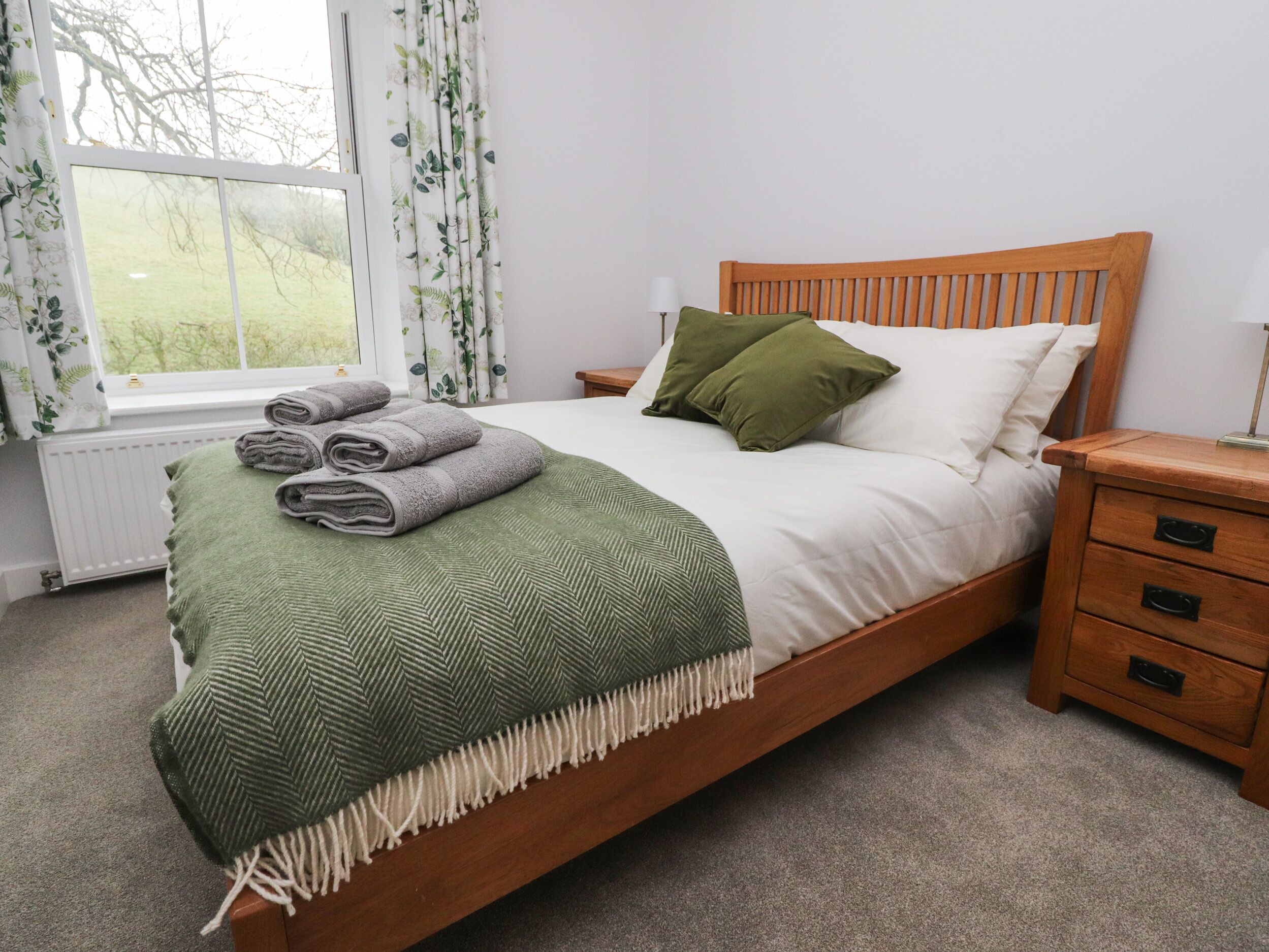A neatly made bed with a green throw blanket and rolled towels, a wooden headboard, a nightstand with drawers, and a window with curtains.