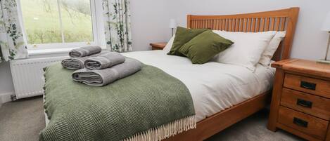 A neatly made bed with a green throw blanket and rolled towels, a wooden headboard, a nightstand with drawers, and a window with curtains.